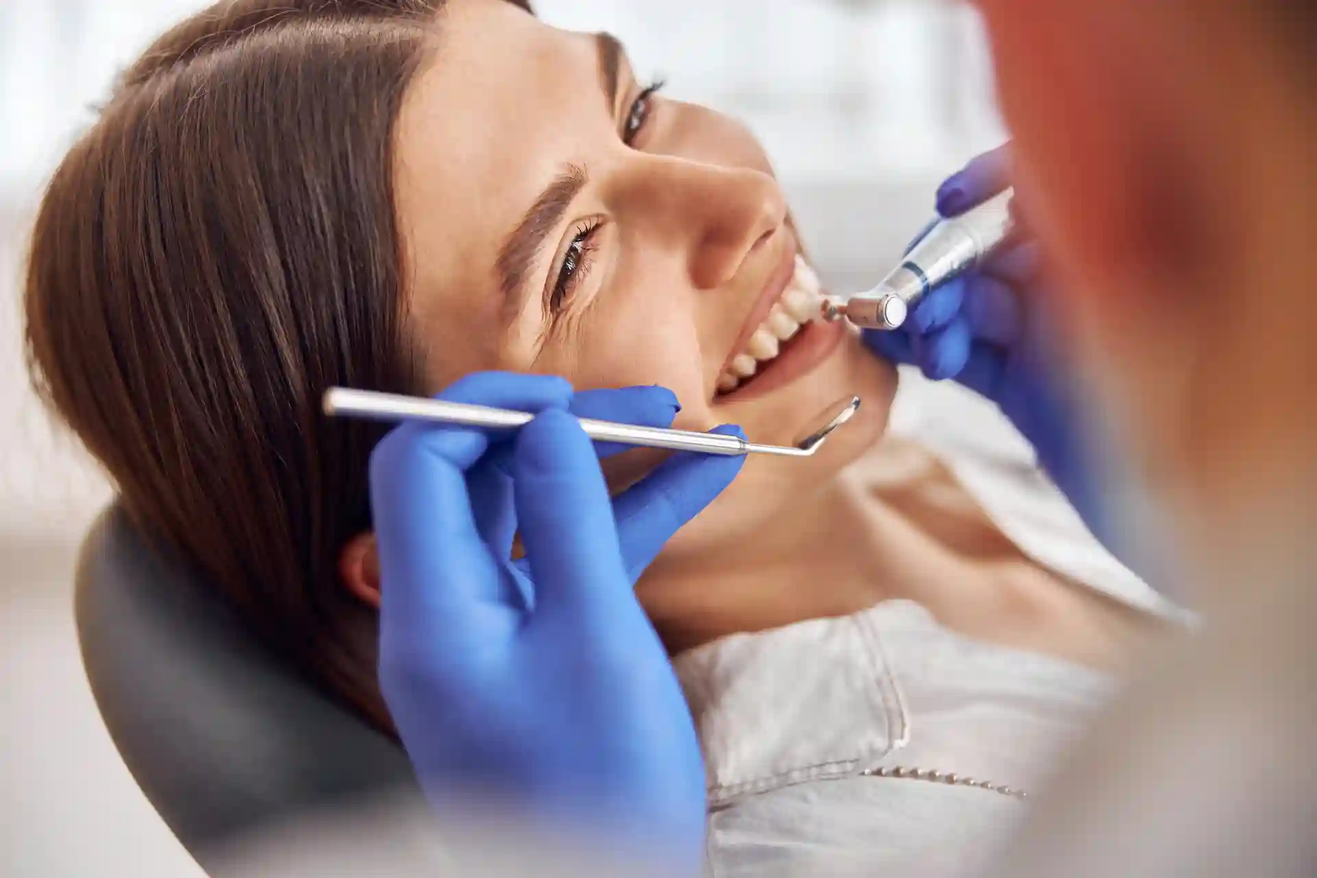 Dentist in blue gloves polishing a woman's upper teeth with a handpiece and mouth mirror in a dental chair