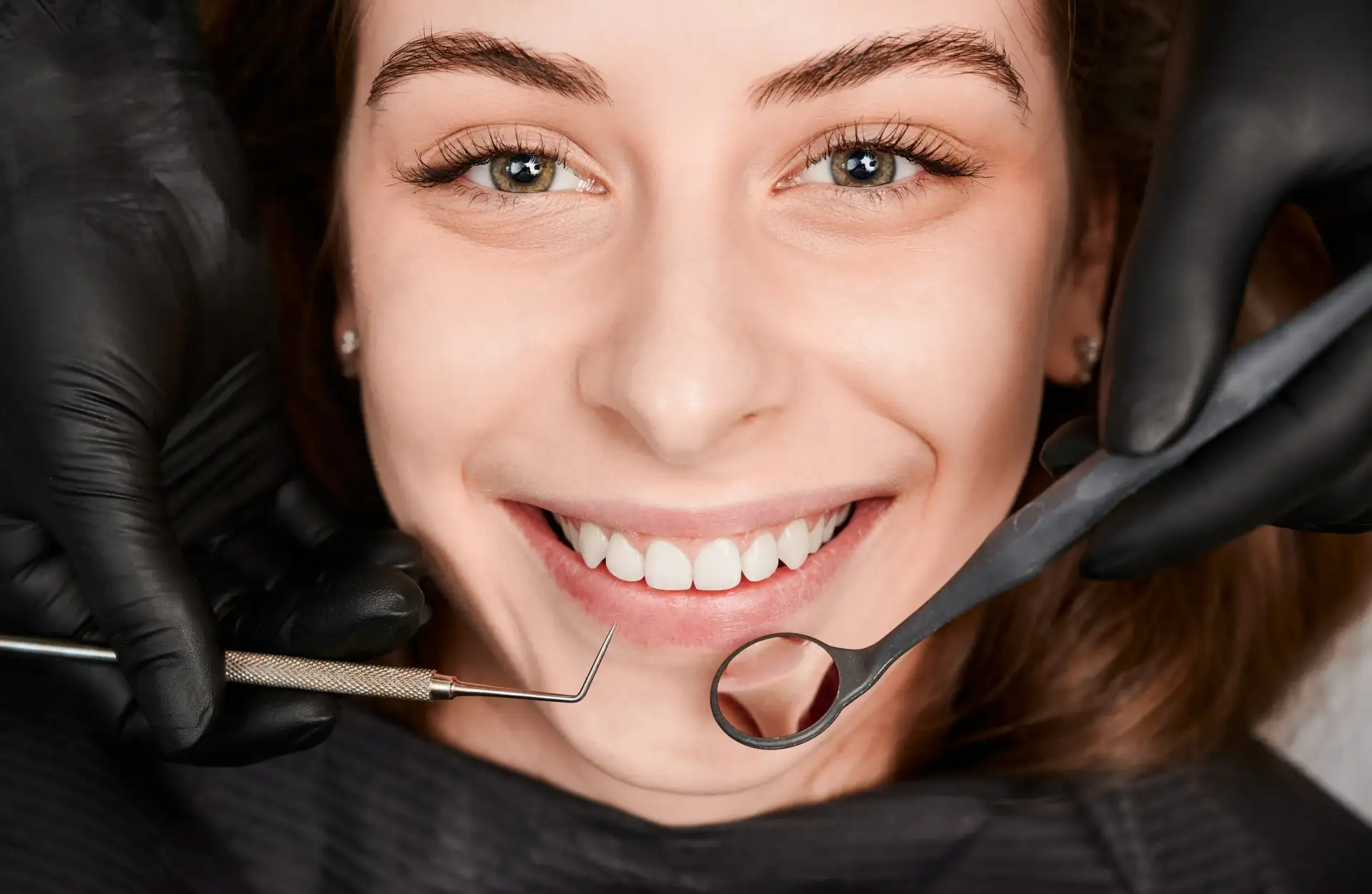 Close-up of a smiling patient in a dental chair while a clinician in black gloves holds a mouth mirror and probe near the upper front teeth.